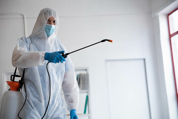 Sanitation worker in protective suit and mask spraying disinfectant under red trim window in room