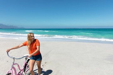 Standing woman wearing orange T-shirt and shorts beside pink cruiser bicycle on sandy beach