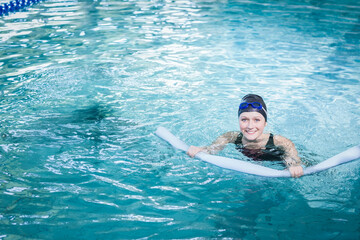 Woman floating in clear blue pool wearing black swimsuit swim cap gripping foam noodle, copy space