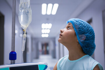 Female child wearing gown and cap standing in hospital corridor observing pump's IV bag, copy space