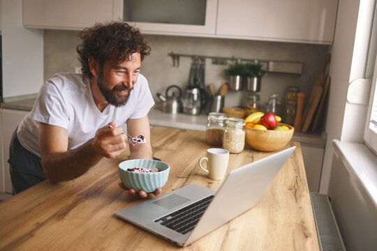 A man is smiling as he eats from a bowl of cereal while seated at a wooden kitchen counter. The laptop is open, and breakfast items are visible in the background.