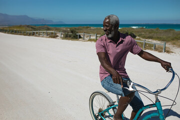 Senior African American man riding teal cruiser bike along coastal path past rail fence, copy space