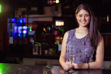Woman in her twenties holding champagne flute behind bar counter at bar lounge, copy space