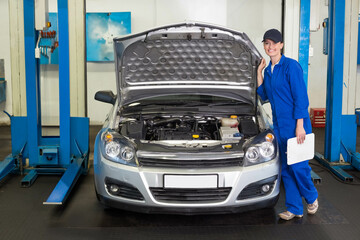 Female auto tech with clipboard inspecting engine under hood open on silver sedan in repair bay