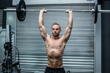 Shirtless man in black athletic shorts lifting heavy barbell overhead on rubber flooring inside gym