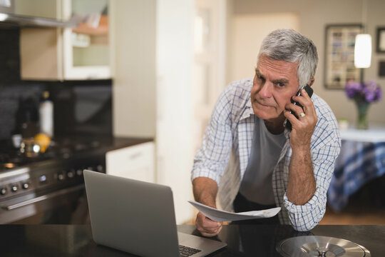 Senior man leaning over kitchen island balancing papers and talking on smartphone by open laptop