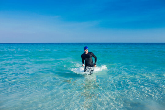 Fototapeta Man emerging from turquoise sea water wearing black wetsuit, blue swim cap, goggles under vivid sky