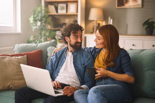 Two people are sitting closely on a sofa in a cozy living room, engaging with a laptop and sharing a warm moment together with smiles and conversation.