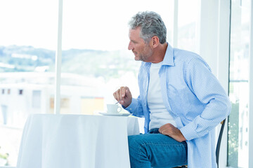 Middle-aged man stirring coffee with teaspoon, cup on saucer at cafe by large windows, copy space