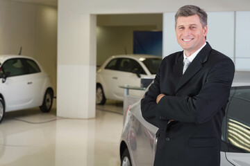Male salesman standing near compact cars with charging cables grey sedan in showroom, copy space