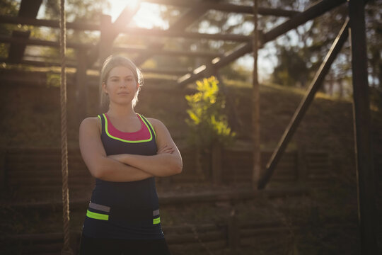 Woman crossing arms before wooden obstacle course structure on wooded hillside in golden sunlight