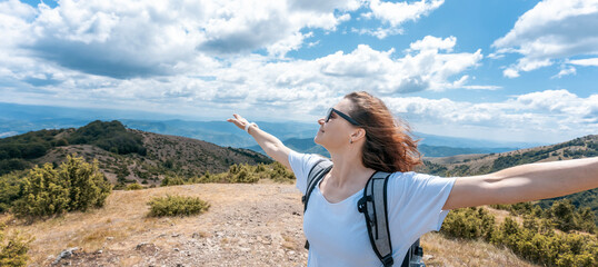 Young happy woman traveler hiker standing with outstretched arms against mountain summer landscape on top