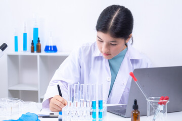 Asian female student scientist in lab coat types on laptop in laboratory while test tubes and...