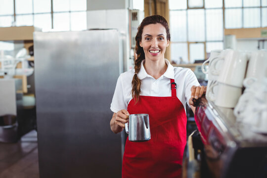 Smiling female barista wearing red apron holding milk pitcher at espresso machine at cafe counter