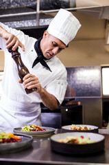 Male chef wearing uniform leaning on counter seasoning plates with wooden pepper grinder in kitchen
