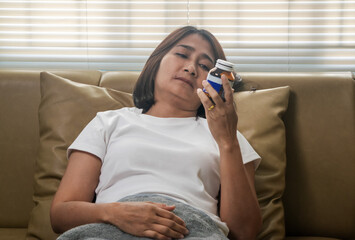 Asian woman lie down on sofa holding bottle reading prescription package  bottle searching pill...