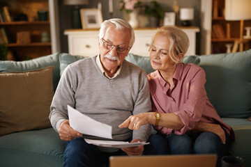 An elderly couple, sitting on a sofa, reviews paperwork and a laptop. The man holds the documents...