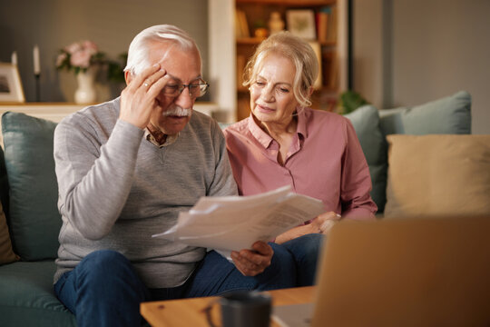 An elderly man and woman are sitting together on a sofa inside their home. They are looking at documents and appear concerned about their finances, possibly related to life insurance.
