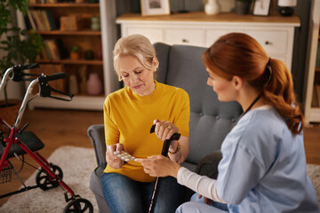 An elderly woman sits in her living room with a red haired caregiver who shows her pills. The...