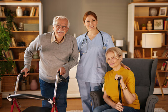 A female home nurse stands behind a senior man using a walker and a seated senior woman holding a cane.