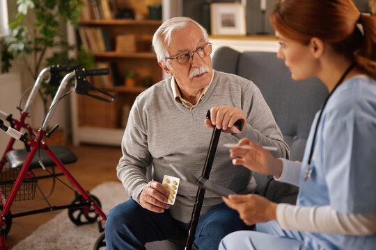 A female home nurse reviews medication with an older man in his living room. He is holding pills and a cane, with a walker nearby, showing he receives assistance with mobility.