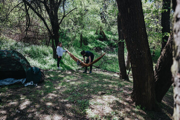 A young couple arranging a hammock between trees in a green forest, with a tent nearby, during a...
