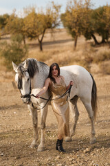 Attractive Caucasian woman in an elegant gold dress leaning on a white horse while holding the...