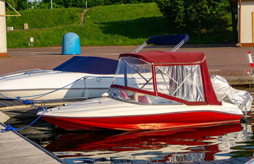 Moored motorboat with a red hull and canopy, reflected in calm water by the quay. Greenery and a...