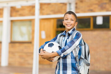 Schoolboy is standing in front of brick school building, holding soccer ball and wearing backpack © wavebreak3
