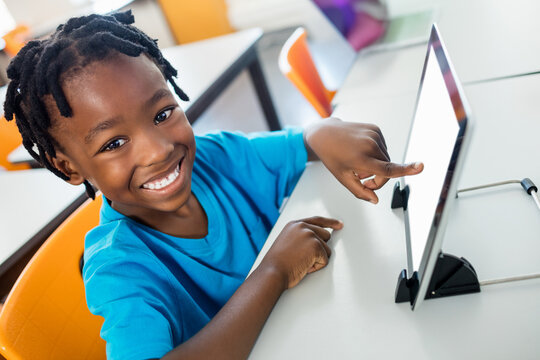 Cheerful African American boy sitting at desk in classroom, pointing at white tablet on black stand