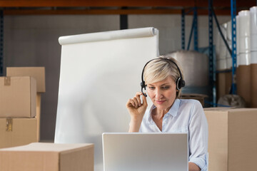 Mature adult woman sitting on cardboard boxes using headset and laptop near flip chart in warehouse