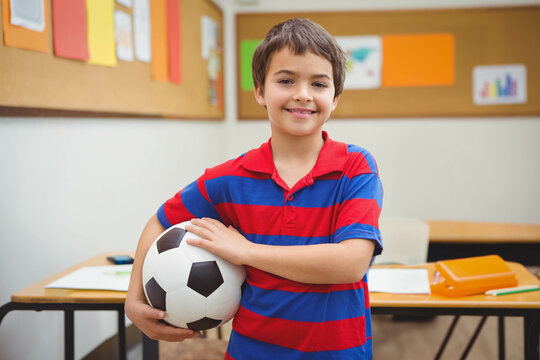 Smiling school-age boy holding soccer ball in classroom among desks, pencil case and bulletin board