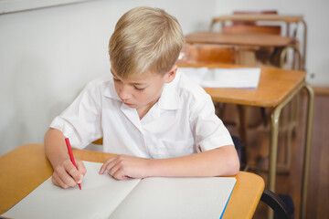 Blonde haired boy sitting at wooden desk in classroom writing in notebook with red pencil