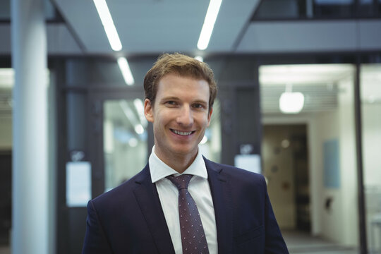 Businessman wearing navy suit jacket and burgundy tie standing under LED lights in office corridor