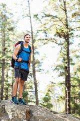 Male hiker standing atop rock outcrop in pine forest with backpack trekking pole and trail shoes