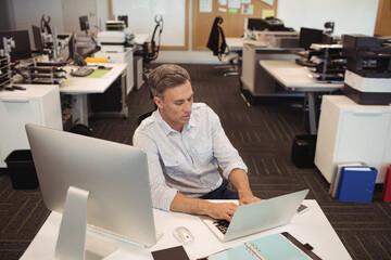 Middle-aged man in business casual attire typing on laptop at desk in open-plan office with monitor