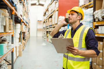 Man wearing vest and helmet checking shelves in warehouse aisle with clipboard, phone, copy space