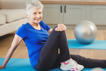 Senior woman twisting torso on blue exercise mat against gray cabinet with silver ball and sofa