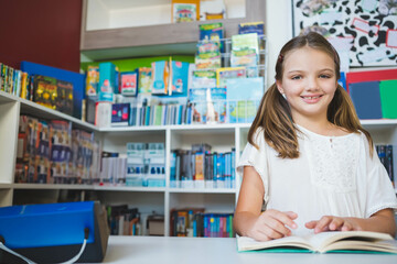 Female child sitting at library desk reading open book with white shelves, blue device, copy space
