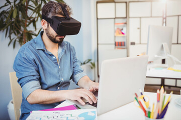 Man wearing black VR headset typing on laptop with sketch papers and colored pencils at desk
