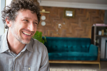 Middle-aged man smiling and sitting in living room with teal sofa, potted plant and shelving unit