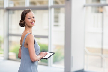Female professional in blue dress holding tablet while looking back in office corridor, copy space