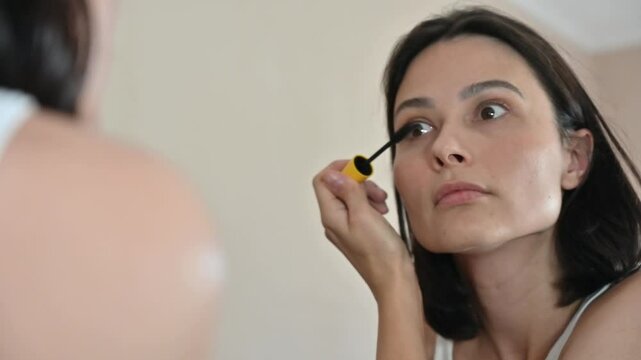 A young brunette woman with a bob haircut carefully applying mascara to her eyelashes in front of a mirror.