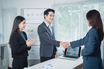 Two asian businesspeople shaking hands in agreement during a meeting, with a colleague clapping her hands in appreciation, in an office environment with charts and a laptop on the table