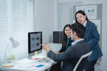 Three asian businesspeople discussing company strategy looking at computer screen showing charts and graphs, working together in modern office with window blinds and lamp