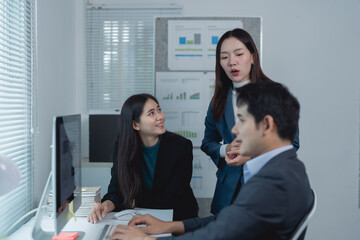 Three asian businesspeople are discussing a project together using a desktop computer in a modern office, with charts and graphs visible on the wall behind them