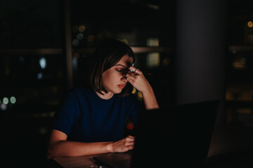 A businesswoman sits at her desk, looking tired and stressed, working on her laptop late at night...