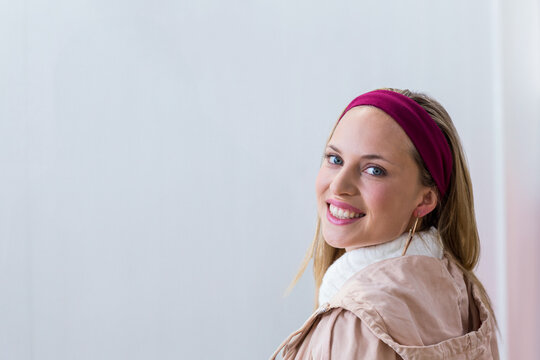Woman wearing magenta headband sweater and khaki jacket smiling and glancing back against pale wall