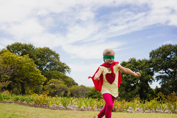 Cape-clad runner wearing DIY superhero costume dashing across manicured park lawn under blue sky