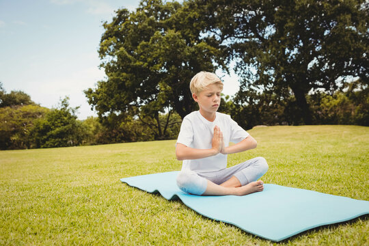 Boy child sitting cross-legged on light blue yoga mat outdoors meditating in grassy park - Powered by Adobe